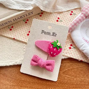 Détail d'une barrette fraise en feutre et d'un nœud rose à rayures sur carte support