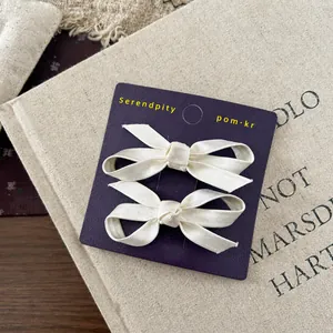 Close-up of a pair of beige ribbon bow hair clips on a purple display card packaging
