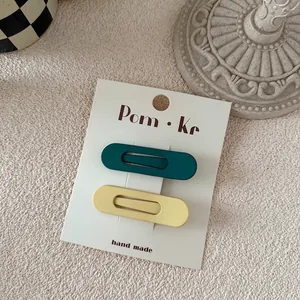 Close-up of two matte oval BB hair clips in dark green and pastel yellow on a paper card