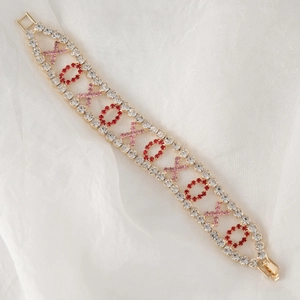 Detail van de goudkleurige XOXO-armband met roze en rode steentjes op witte stof