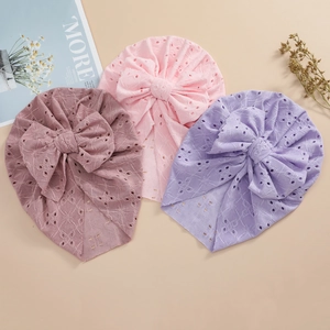 Pink and purple hollow knit baby turban beanies with large bows displayed on a beige surface