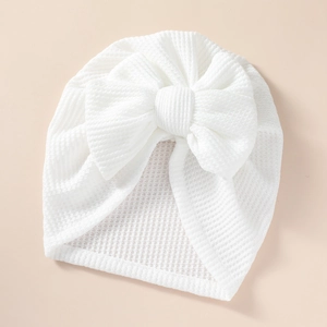Close-up of white waffle knit baby turban hat with a large decorative bow on light background