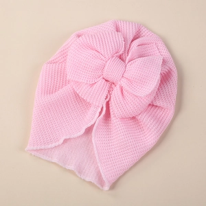 Pink knit baby turban hat with large double bow decoration and wavy edges on beige background