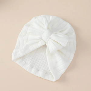 Detail of white knitted baby turban hat featuring a large soft bow on a neutral background