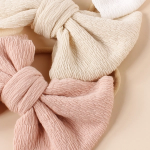 Three soft knit baby headbands with large textured bows in pink, beige, and white colors