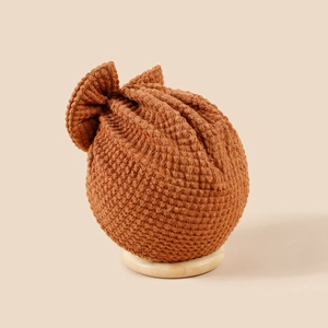 Bonnet turban en tricot pour bébé couleur caramel avec grand nœud sur socle bois