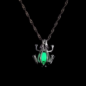 Detailed close-up of a silver frog pendant glowing green in the dark on a black background