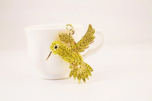 Détail du pendentif colibri avec strass jaunes exposé sur une tasse blanche