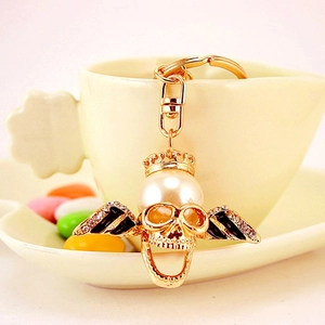Golden skull keychain with pearl crown and rhinestone wings on a beige cup