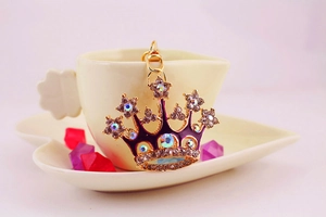 Detail of a gold crown keychain with purple enamel and diamonds hanging on a white coffee cup