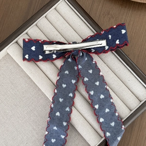 Navy denim-style bow hair clip with white hearts and red stitching on a jewelry tray