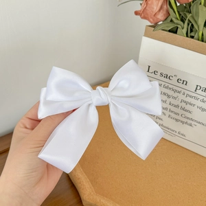 Detail view of a creamy white satin bow hair clip held by hand in a minimalist setting