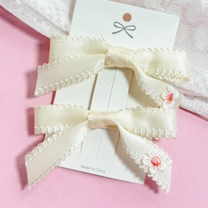 Pair of cream bow hair clips with delicate floral embroidery and lace trim on pink background