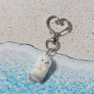 Cute plush sea otter keychain with silver heart clasp on a sandy beach background