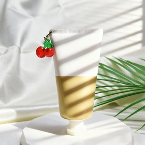 Minimalistische Kosmetiktube in Weiß und Gold mit Kirsch-Anhänger und grünen Blättern