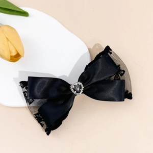 Détail d'une pince à cheveux nœud en dentelle noire avec gemme cœur sur plateau blanc
