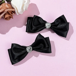 Detail von zwei schwarzen Satin-Schleifen-Haarspangen mit Herz-Strass-Dekor auf rosa Hintergrund