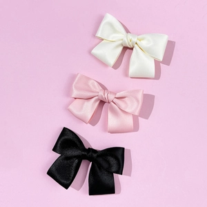 Three-piece set of satin bow hair clips in cream, pink, and black on a purple background