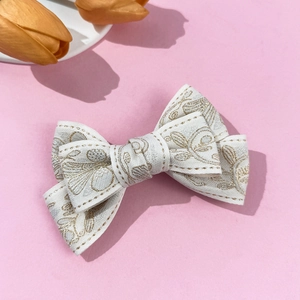 Elegant beige bow hair clip with golden flower embroidery on a soft pink background