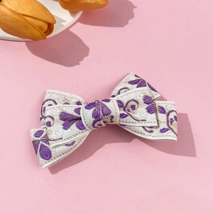 Detailed close-up of white fabric bow hair clip with purple floral embroidery and gold thread