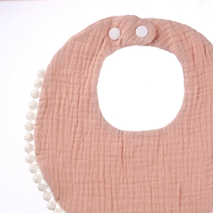 Close-up of pink muslin baby bib featuring white pom-pom trim and adjustable snap buttons