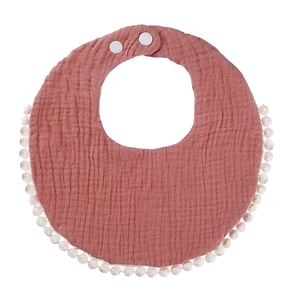 Detail view of terracotta cotton muslin baby bib with white pom-pom trim and snap buttons