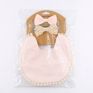 Pink baby bib and headband set with beige pompom trim and lace bow in packaging