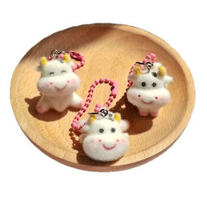 Three cute flocked cow plush keychains on a wooden tray with pink beaded chains