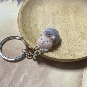 Miniature plush hedgehog keychain with fluffy pink face and grey body on a wooden tray