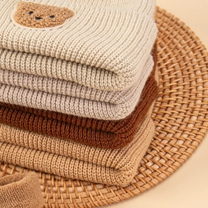 Detail of ribbed knit baby hats with bear patch on a rustic woven mat in warm neutral tones