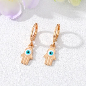 Golden Hamsa Hand earrings with Evil Eye detail on a clean white stone surface