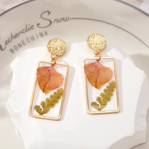 Macro detail of rectangular gold earrings with preserved pink dried flowers and green fern leaf