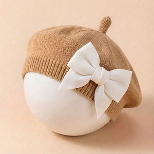 Détail d'un béret tricoté pour bébé avec nœud blanc sur tissu beige, vue de près