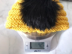 Detailed weight measurement showing light yellow knit baby hat with black pompom on a digital scale