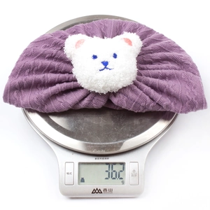 Detail view of purple knitted baby headband with bear decor on a scale weighing 36.2g