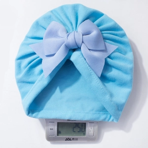 Détail montrant le bonnet turban bébé de 25g avec un grand nœud violet sur balance