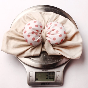 Detailed view of lightweight 30g beige baby turban with red polka dot bow on a scale
