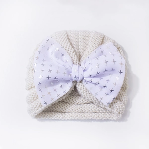 Close-up of knit baby beanie featuring a large silver-patterned white bow on beige fabric