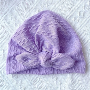 Detail of lilac knitted baby hat with ribbed texture and decorative bow on white background
