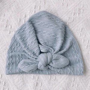 Light blue cable knit baby turban hat with a large decorative bow on a white textured background