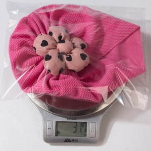 Pink infant flower turban hat weight shown on a kitchen scale in packaging