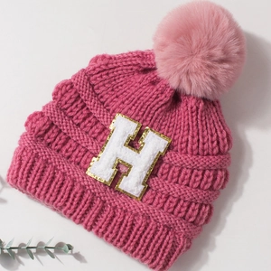 Detail of rose red knit hat featuring a sequined H letter patch and pink faux fur pompom
