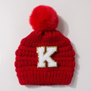Detailed view of red knitted baby hat with fluffy pompom and gold embroidered letter K
