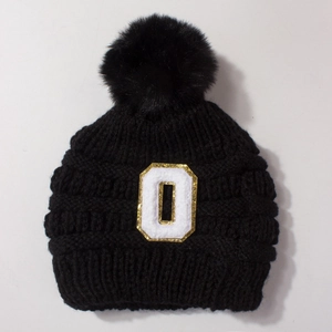 Close-up of black knitted baby hat with large fur pompom and gold sequin letter O patch
