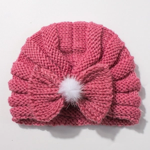 Detail of rose pink cable knit baby hat featuring large bow and fluffy white pom-pom decoration