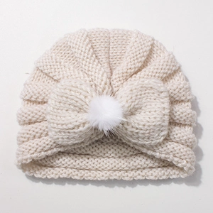 Detail of a beige knit baby turban hat featuring a large soft bow and faux fur pompom