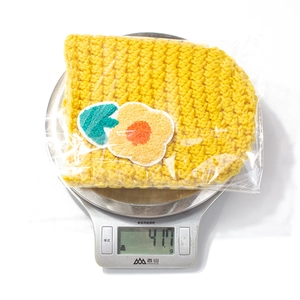 Detailed view of yellow knit baby hat with flower patch being weighed at 41.7 grams