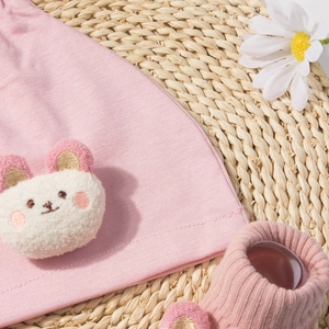 Pink modal baby hat and socks set with a white plush bear patch on woven mat