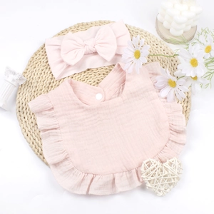 Pink cotton gauze baby bib with ruffle edges and matching bow headband on a straw mat