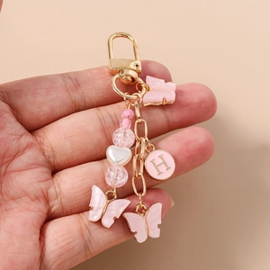 Porte-clés papillon rose avec lettre H, perles et fermoir doré tenu dans la main d'une femme